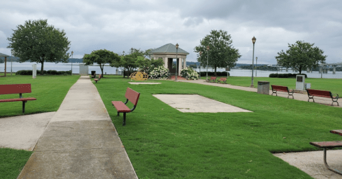 A park with green grass, benches, a gazebo, and a view of a lake under a cloudy sky.