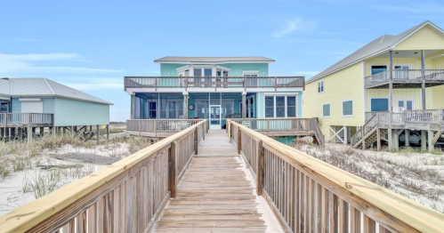 A wooden walkway leads to a beach house, flanked by two colorful homes, with sandy dunes in the foreground.