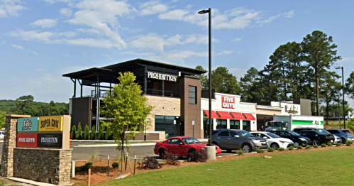 A modern building with multiple restaurants, including Prohibition and Five Guys, surrounded by parking and greenery.