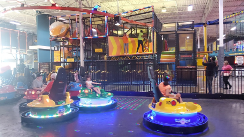Children enjoy bumper cars in a vibrant indoor amusement center with a climbing structure in the background.