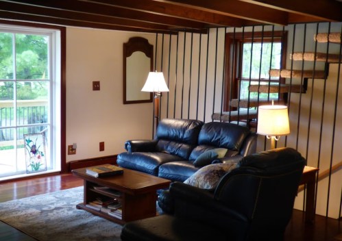 Cozy living room with a blue leather sofa, wooden coffee table, and a staircase, featuring large windows and warm lighting.
