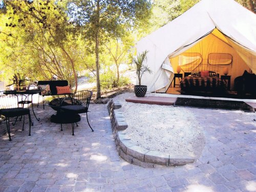 A cozy glamping tent surrounded by trees, with outdoor seating and a stone pathway.