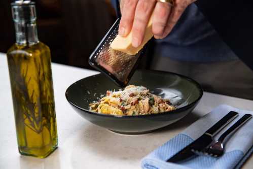 A hand grates cheese over a bowl of pasta, with a bottle of olive oil and utensils nearby on a table.