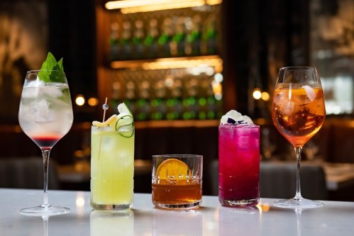 Five colorful cocktails displayed on a bar counter, with a blurred background of bottles and warm lighting.