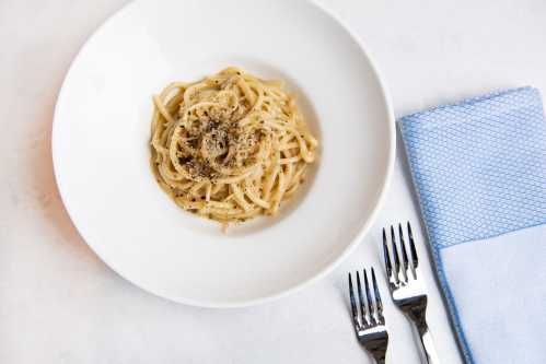 A bowl of spaghetti topped with black pepper, served with two forks and a blue napkin on a white surface.