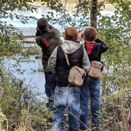 Three children with backpacks observe a body of water through trees, capturing the scene with their cameras.