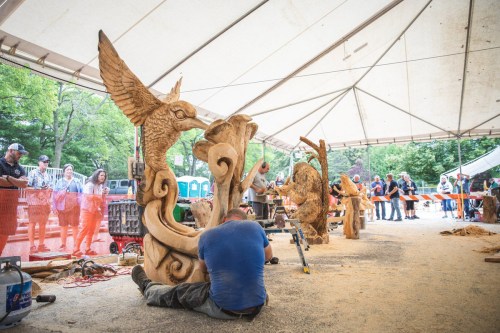 A woodcarver works on a sculpture under a tent, with spectators observing and other sculptures displayed nearby.