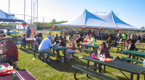 A lively outdoor gathering with people dining at picnic tables under a large tent, decorated with colorful balloons.