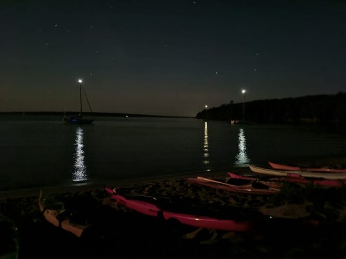 A serene night scene at a beach with kayaks on the sand, calm water reflecting lights from boats and stars above.