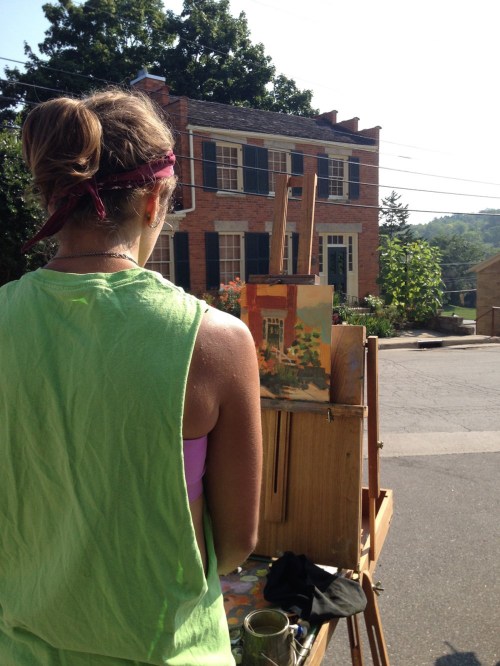 A person in a green tank top paints on an easel, facing a brick house with green shutters in the background.