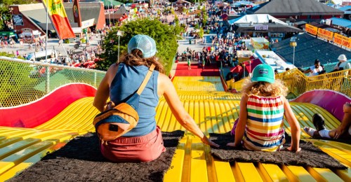 A woman and a child sit on a colorful slide, overlooking a busy fairground filled with people and attractions.