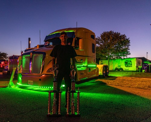 A person stands with trophies in front of a brightly lit truck at dusk, showcasing vibrant green lights.