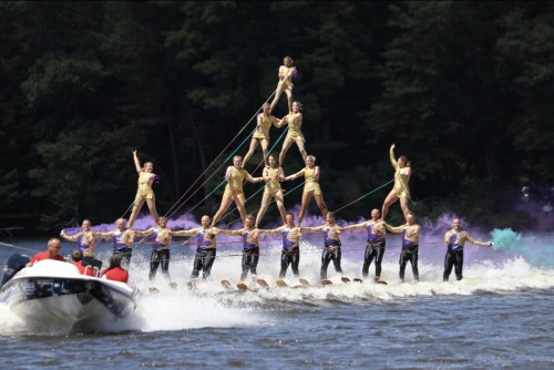 A team performs a pyramid formation on water skis, with colorful smoke in the background and a boat nearby.