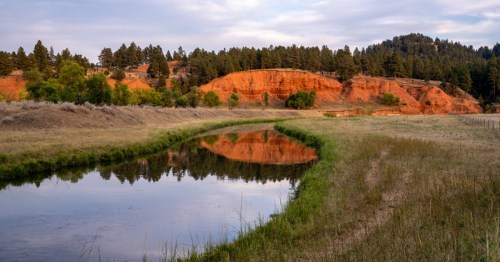 A serene river reflects red rock formations and green trees under a cloudy sky in a tranquil landscape.