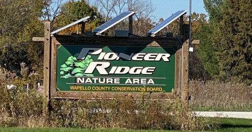 Sign for Pioneer Ridge Nature Area, featuring solar panels, with greenery and trees in the background.