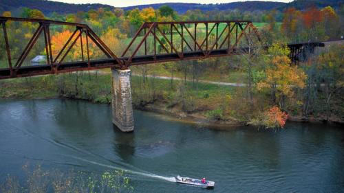 A boat glides on a river beneath a rusted train bridge, surrounded by colorful autumn foliage.