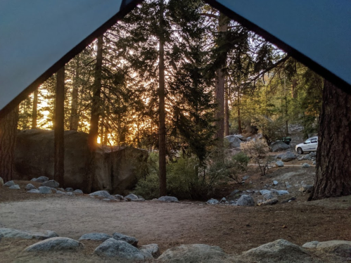 View from a tent showing a sunset through trees, with rocks and a parked car in the background.