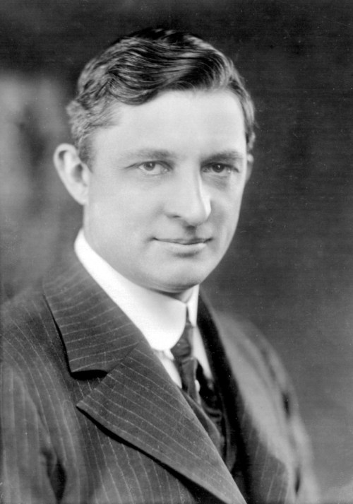 A black and white portrait of a man in a suit, with short wavy hair and a serious expression.