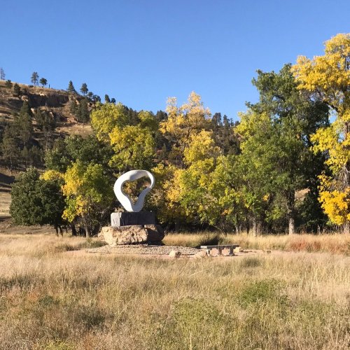 A large abstract sculpture on a stone base surrounded by colorful trees and grassy land under a clear blue sky.