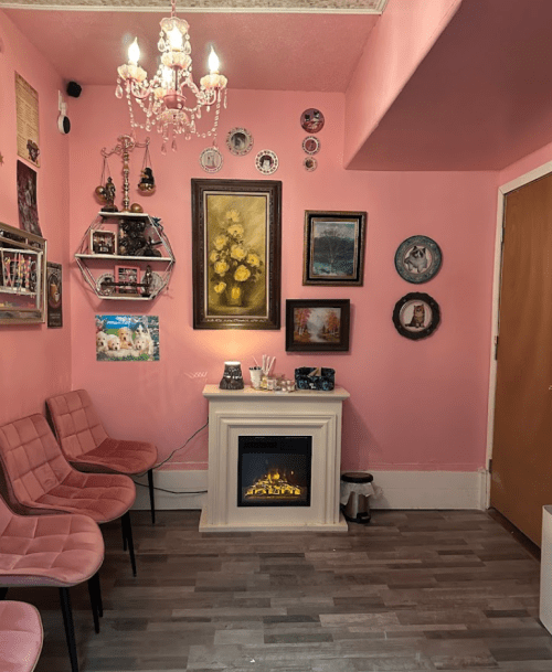 Cozy pink room with a chandelier, decorative wall art, and a faux fireplace surrounded by stylish chairs.