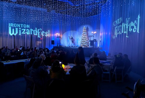A festive event scene with a stage, Christmas tree, and audience under twinkling lights, featuring "Ironton WizardFest" signage.