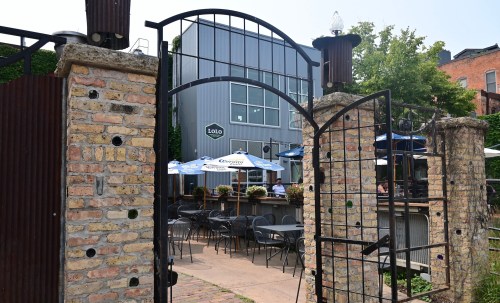 A gated entrance leads to a patio area with tables and umbrellas, surrounded by brick walls and greenery.