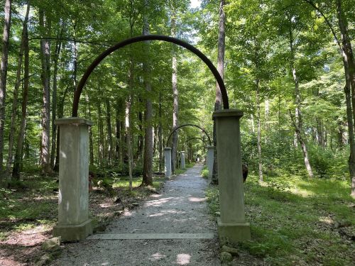 A serene forest path lined with stone pillars and arches, surrounded by lush green trees and dappled sunlight.