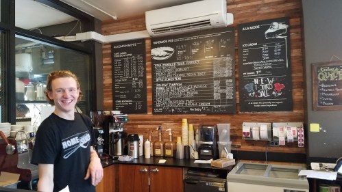 A smiling barista stands behind a coffee counter with a menu board and various coffee supplies in a cozy café.