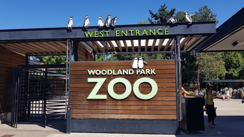 West entrance of Woodland Park Zoo, featuring a wooden sign and penguin decorations on top. Visitors are entering.