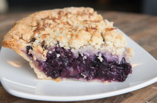 A slice of blueberry pie with a crumbly topping on a white plate, resting on a wooden table.