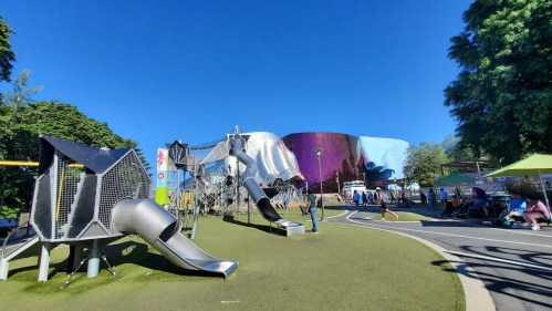 A vibrant playground with slides and climbing structures, surrounded by greenery and a colorful building in the background.