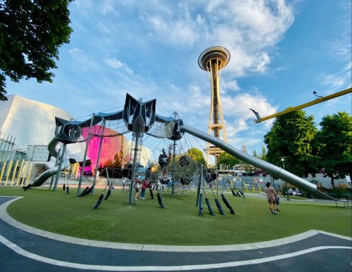 A playground with a large climbing structure, the Space Needle in the background, and people enjoying the park.