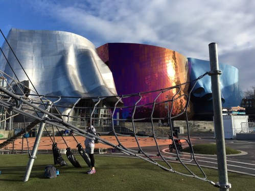 A modern building with colorful, metallic surfaces, viewed through a playground's climbing structure.