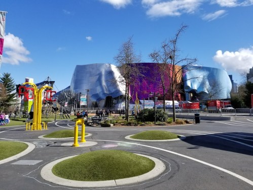 A modern building with colorful, metallic surfaces, surrounded by trees and a park area under a clear blue sky.