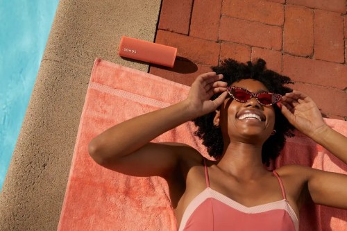 A woman in sunglasses relaxes by a pool, smiling on a towel with a portable speaker nearby.