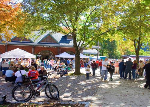 A bustling outdoor market scene with people, tents, and trees in autumn colors. Bikes are parked nearby.