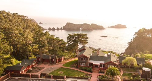 Aerial view of coastal homes surrounded by trees, with rocky islands and ocean in the background during sunset.