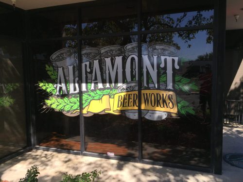 Sign for Altamont Beer Works displayed on a glass window, featuring beer cans and green hops design.