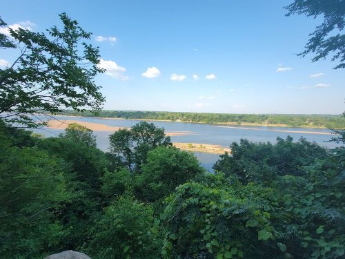 A scenic view of a river surrounded by lush greenery and trees under a clear blue sky with scattered clouds.