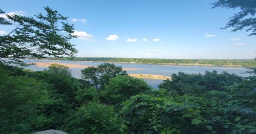 A scenic view of a river surrounded by lush greenery and trees under a clear blue sky with a few clouds.