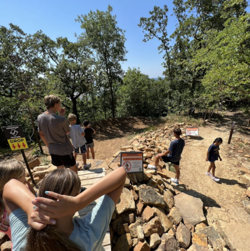 A group of children and teens at a rocky trail with warning signs, surrounded by trees on a sunny day.