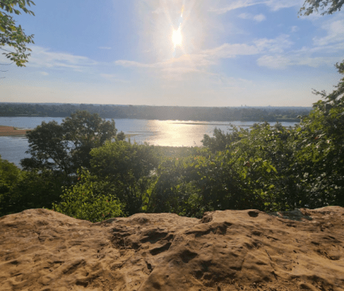 A scenic view of a river reflecting sunlight, surrounded by trees and a rocky foreground under a clear blue sky.