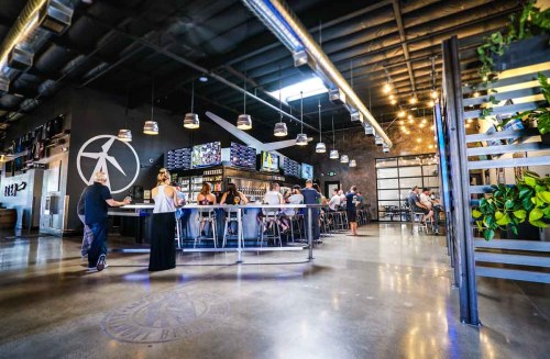 A modern bar interior with patrons at the counter, high ceilings, and stylish decor featuring plants and industrial elements.
