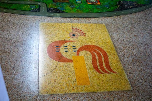 A colorful mosaic of a rooster on a textured floor, featuring vibrant patterns and a cracked surface.