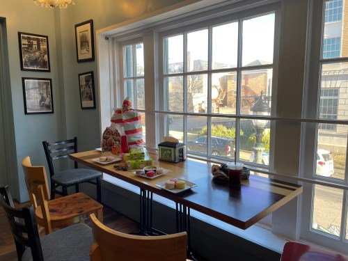 A cozy café scene with a table set by a large window, featuring snacks and drinks, and chairs around it.