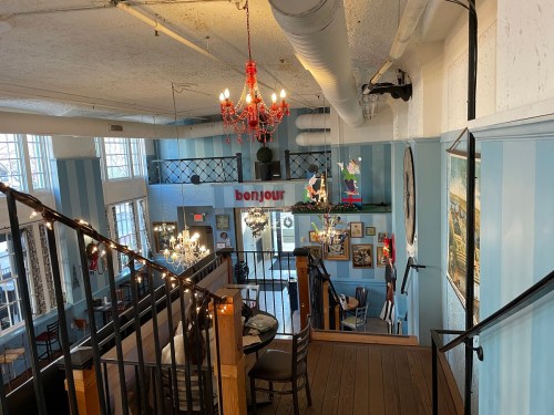 A cozy café interior with blue striped walls, chandeliers, and a welcoming "bonjour" sign. Stairs lead to a second level.