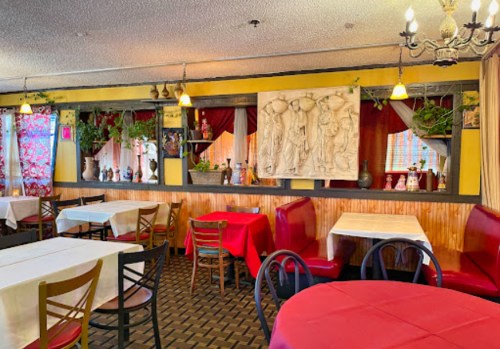 Cozy restaurant interior with red and white tablecloths, decorative wall art, and plants in a warm, inviting atmosphere.