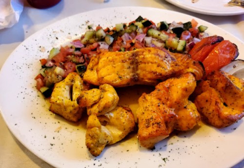 Grilled chicken pieces served with a fresh salad of diced tomatoes, cucumbers, and grilled vegetables on a white plate.