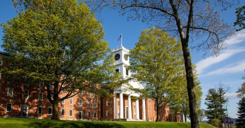 A brick building with a clock tower, surrounded by green trees and a clear blue sky.