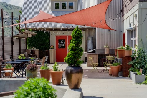 A cozy outdoor patio with potted plants, tables, and a red shade sail, leading to a door with "Eat Well" sign.